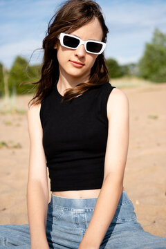 A teenage girl in jeans stands poised in a desert coastal landscape, arms bent and hair caught in the wind. The teenager projects confident stylish attitude against sparse dunes and open sky.