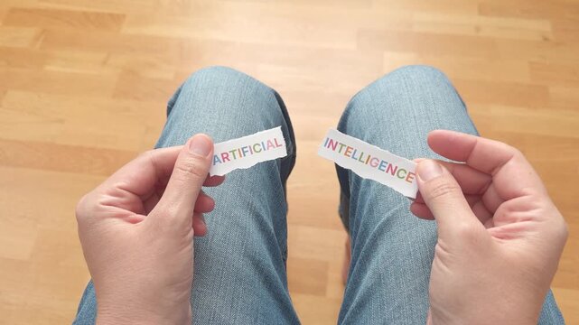First-person point of view of a woman sitting in jeans and tearing a piece of paper with the words Artificial Intelligence printed in colorful letters.