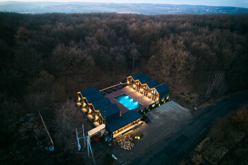Obraz premium Aerial night view shows modern resort cabins around glowing pool. Warm lights, spacious wooden terrace and secluded forest setting create inviting luxury retreat concept
