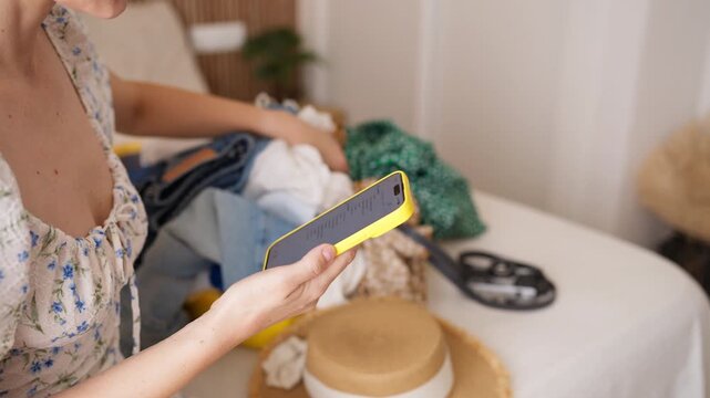 Young woman organizing her summer clothes and accessories for an upcoming vacation, checking a digital packing list on her smartphone while sitting by her suitcase