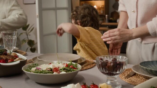 Caucasian child carrying bowl of fresh salad to dining table with Caucasian woman helping him, preparing family dinner. Clip for healthy eating ads