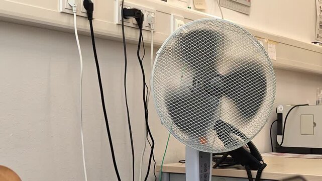 Fan rotating, in indoor space with white walls and ceiling, with fans, wires, with metallic fan blades, plastic fan base texture, from a side view perspective, with white, gray accents.