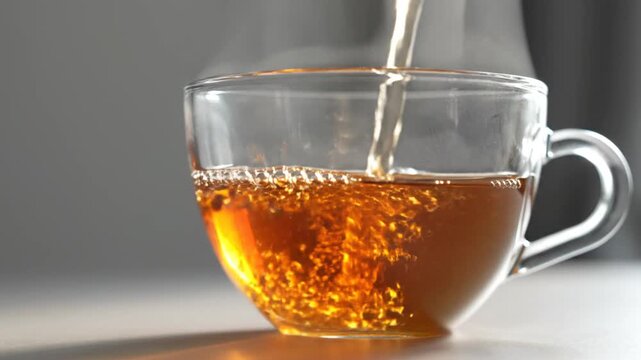 Freshly brewed hot tea being poured into a transparent glass cup, with steam rising and bubbles forming as the liquid fills the vessel in a cozy, brightly lit setting.
