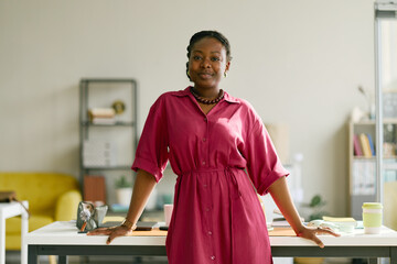 Obraz premium Portrait of young African American businesswoman standing at desk in office, looking at camera and presenting confident leadership. Ideal for workplace branding, career growth campaigns