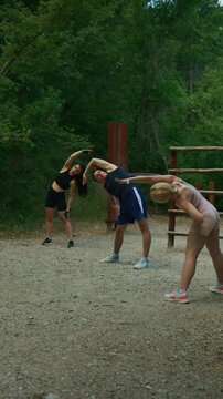 A group of friends is stretching on a gravel path in a lush green park. The young adults are warming up with fitness exercises outdoors.