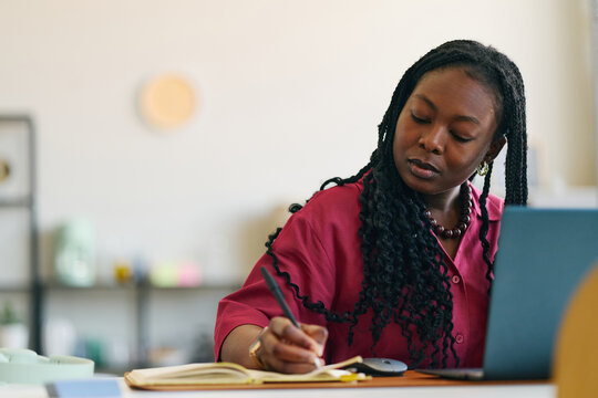 Young African American woman writing notes beside laptop, studying or planning work tasks at desk. Ideal for remote work, online learning, productivity, business content