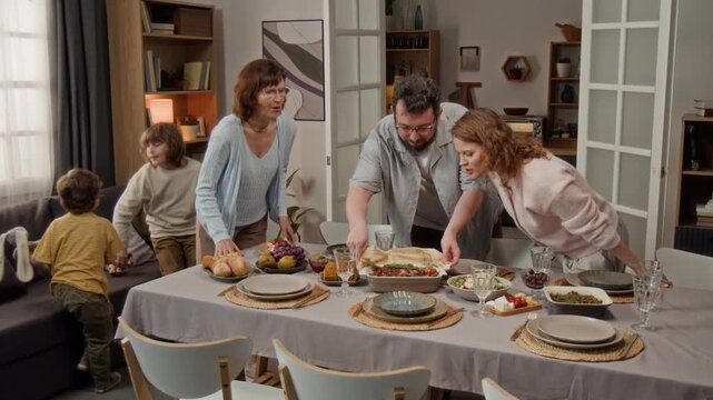 Family preparing dinner table in dining room, man bringing hot dish while women arranging plates and children playing. Perfect for family lifestyle and food marketing campaigns
