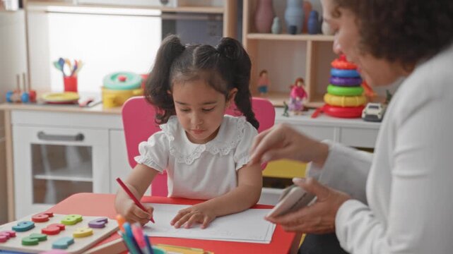 Little girl writing with red pencil on paper at a preschool desk while woman teacher displays a flashcard in a classroom building; concentration learning.