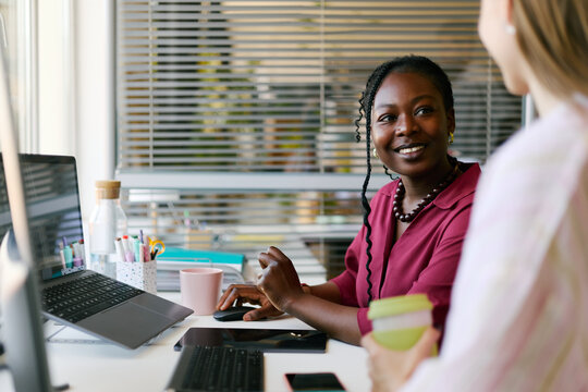 African American young adult businesswoman discussing project with Caucasian colleague while working at desk with laptop, supporting teamwork and office communication for corporate marketing