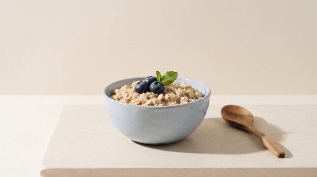 Oatmeal porridge topped with fresh blueberries in ceramic bowl next to wooden spoon in minimalist studio style in light blue and beige tones representing healthy organic breakfast with copy space