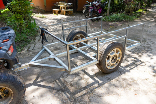 Lightweight metal utility trailer frame with two wheels parked on sunlit driveway, rugged tire rims and open rectangular steel structure ready for cargo hauling