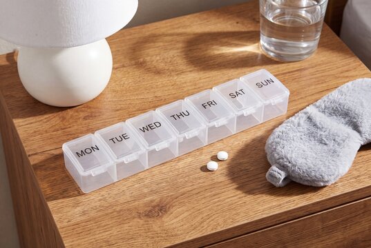 Seven day pill organizer with weekday labels MON through SUN beside fuzzy sleep mask on a wooden nightstand
