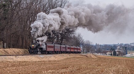 A steam train moves along the tracks in a rural area with fields and trees. Dark smoke billows from the engine as it travels. The weather is cloudy and the scene shows the train making its journey. © Greg Kelton