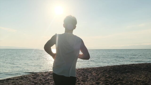 Camera follows the back of a handsome young athlete with muscular arms and flowing hair running along the seashore at golden sunset. Morning run, healthy lifestyle, determined runner chasing records.