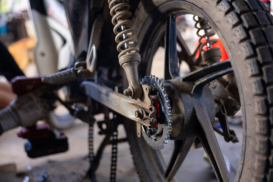Rear motorcycle wheel and sprocket close up showing metal chainring, suspension spring and worn tire with oil stained swingarm and workshop tools, gritty mechanical mood