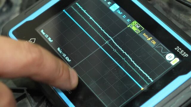 A close-up of a person's finger touching a portable digital oscilloscope screen showing waveforms.
