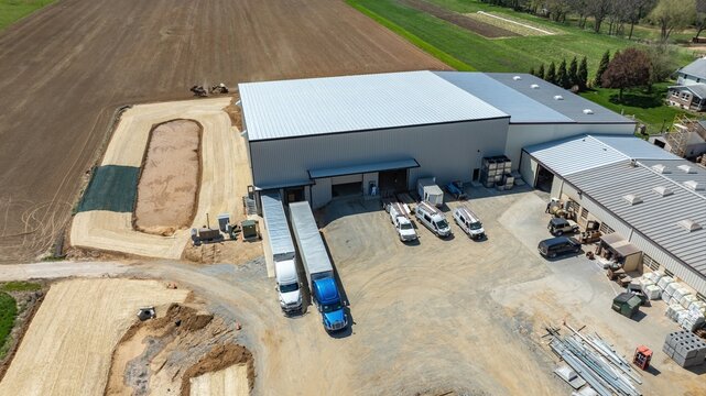 Workers are present at a construction site with trucks parked outside a large building. Machinery is near the site, and the surrounding area shows fields and dirt roads.