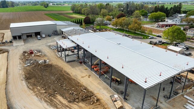 Workers are constructing a large building at a construction site in a rural area. Heavy machinery and vehicles are present. Green fields and trees surround the location, showing a peaceful landscape.