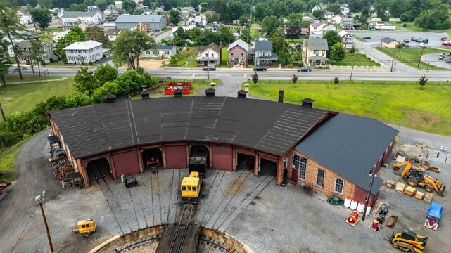 A steam engine roundhouse is seen from above in a small town. Various buildings surround the structure. Equipment and vehicles are parked outside.