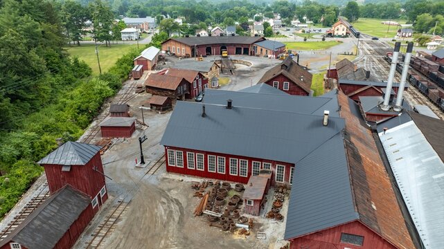 An overview of a historical train yard with multiple red workshop buildings and a simple landscape. The area is surrounded by trees and some small town buildings are nearby.