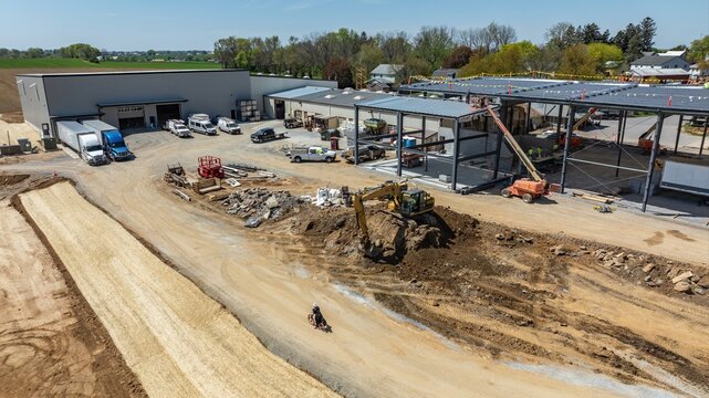 Construction is taking place at a site in a rural area. Workers and machinery are present. Trucks are parked nearby. The ground is uneven with dirt and building materials scattered around.