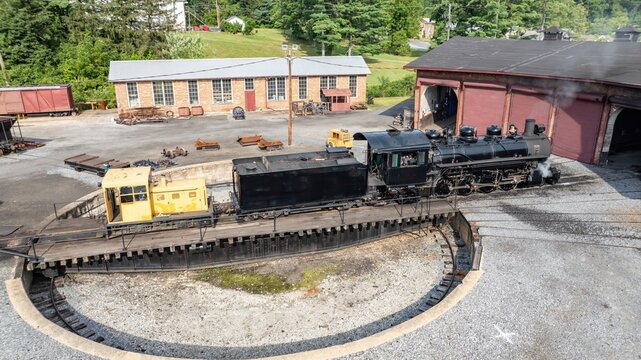 A steam engine is on a turntable at a railway work site during the day. This site has buildings and tracks nearby. Workers might perform maintenance tasks here.