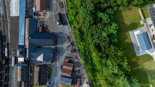 An old industrial area next to a green space in a town. The image captures buildings and a forested area. Nearby, there is an open field and some structures visible.