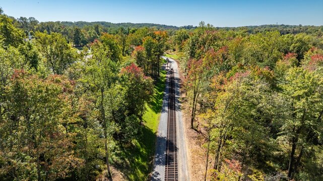 A view shows train tracks stretching through a forested area. Trees with changing leaves surround the tracks on a clear day. The scene is calm with blue sky above.