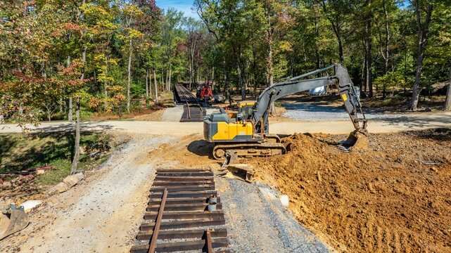 Construction is taking place in a rural area. An excavator moves dirt and debris while another machine is visible in the background. Trees surround the worksite under clear skies.