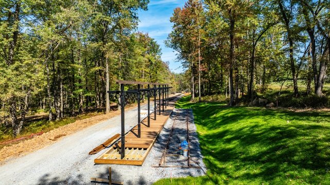Workers are building a wooden deck along a gravel path in a forest. Trees surround the area, and the sky is clear. Tools and materials are scattered around as work is in progress.