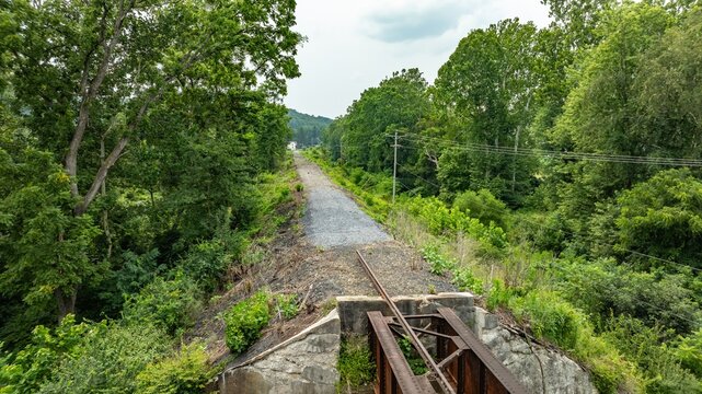 An abandoned railway track stretches into the distance through a green area. Trees line both sides, and the track seems unused. The scene is set in a rural location on a clear summer day.