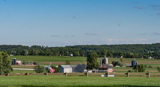 Fields and farms spread across the landscape under a clear sky. Red barns and silos stand among green trees. An American flag waves gently by the wooden fence in the foreground.