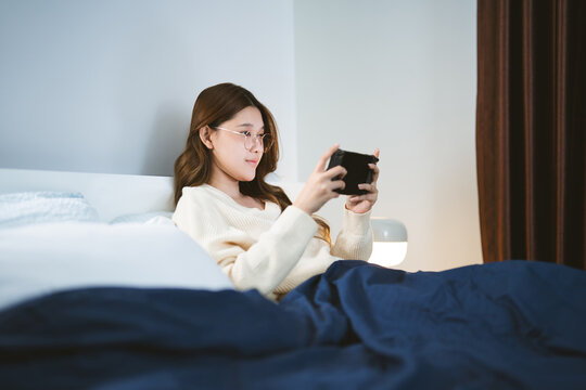 Happy smile young adult asian gamer woman wear eyeglasses play a online game portable handheld in bedroom at night
