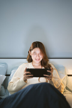 Happy smile young adult asian gamer woman wear eyeglasses play a online game portable handheld in bedroom at night
