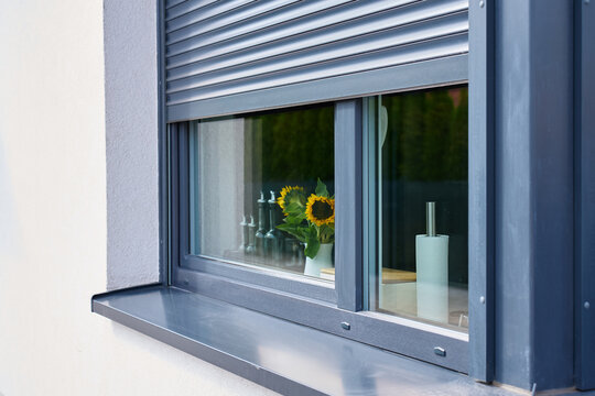 Partially closed exterior roller shutter shades kitchen window with sunflowers and glass bottles inside