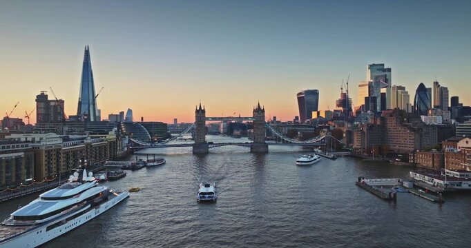 Amazing aerial view of iconic London skyline featuring The Shard, Tower Bridge, boats navigation Thames River, and modern City of London skyscrapers glistening during a beautiful sunset. Drone flight
