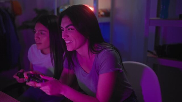 Two women holding game controllers, hands gripping joysticks, smiling and leaning forward while playing in a studio with neon shelving and lamp; excitement friendship.