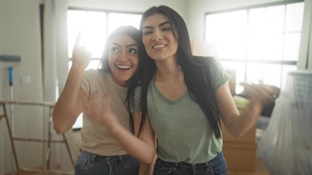 Two women smiling and waving hands while standing by cardboard boxes and a ladder in an apartment building, casual jeans and tshirts visible; friendship joy.
