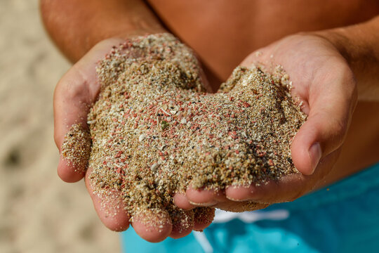Cupped hands holding a heap of pink shell sand on the beach, turquoise swim shorts behind, Voidokilia beach, Greece