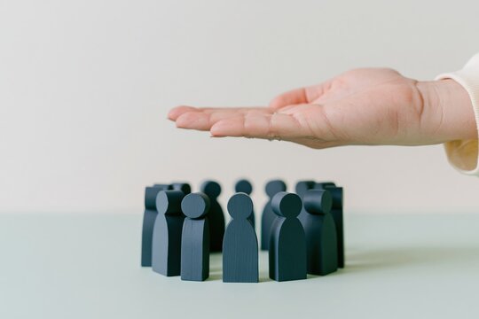 A womans hand cupped protectively over blue wooden figurines arranged in a circle, representing a soft and caring community for humanism and protection