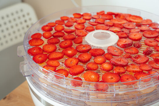 Detailed view of sliced red strawberries on transparent trays of an electric food dehydrator. Healthy snacks drying indoors in a home kitchen environment.