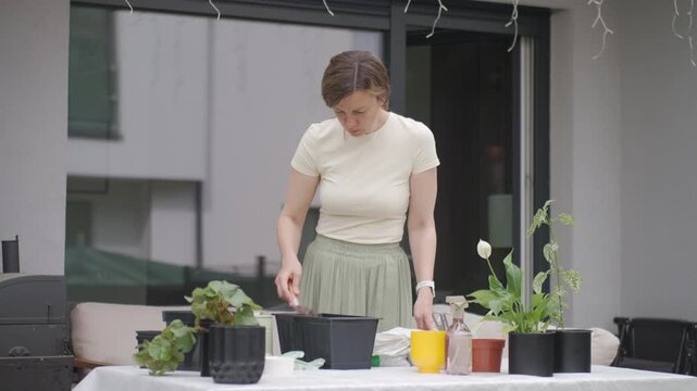 Woman adds clay pebbles to flower pot, filling planter with pink scoop at gardening table. Concept of container gardening and plant drainage