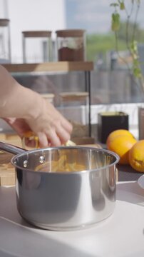 Hand adds orange peel strips to saucepan on kitchen counter. Concept of making candied orange peel and citrus dessert