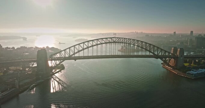 Aerial drone panorama of Sydney Harbour Bridge spanning the tranquil harbor waters with the iconic Opera House visible in hazy morning light, revealing the coastal cityscape and urban infrastructure