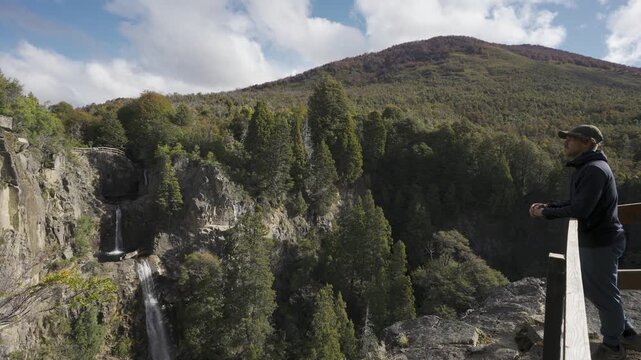 Man standing on a wooden viewpoint looking at Los Cesares waterfall cascading over a rugged cliff in Nahuel Huapi National Park, Rio Negro, Argentina.