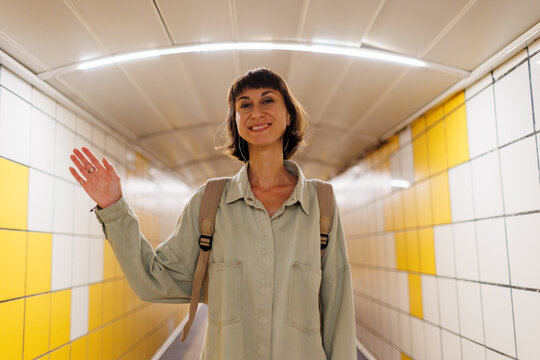 Friendly, cheerful, happy smiling woman with backpack waving at you with raised hand. Attractive girl greeting friend, saying hi or hello, greeting guest.