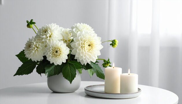 A beautiful bouquet of fresh white dahlia flowers in a small minimalist vase with two lit pillar candles on a table for home decor.