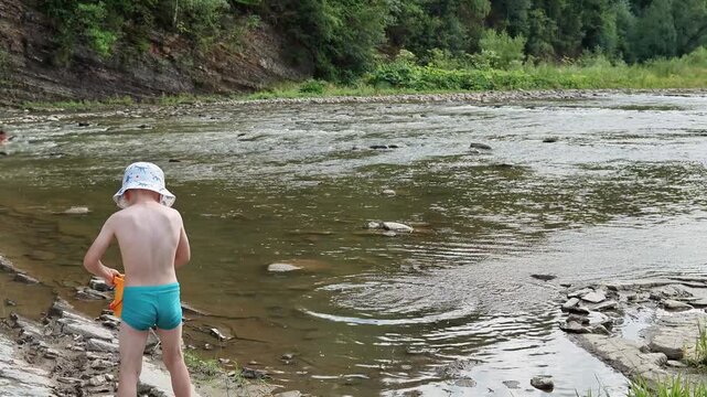 Child boy plays with stones on the river. Takes stones from a bucket and throws them into the water. Vacation, leisure, rest in a mountain river. Happy childhood. slow motion