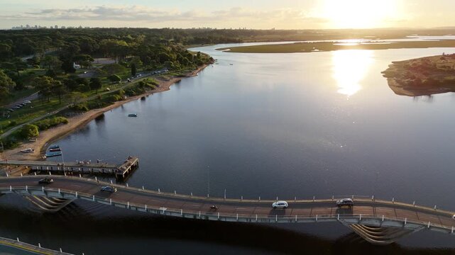 Aerial drone footage of La Barra Bridge crossing the during golden hour in Punta del Este, Uruguay. Scenic coastal lagoon, wetlands, calm water and evening sunlight captured from above