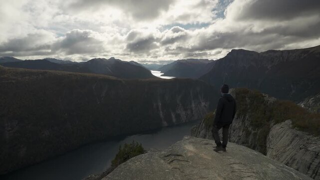 Hiker standing on the edge of a high mountain peak at La Mirada del Doctor, looking out at a reflective lake nestled in Nahuel Huapi National Park, Argentina.